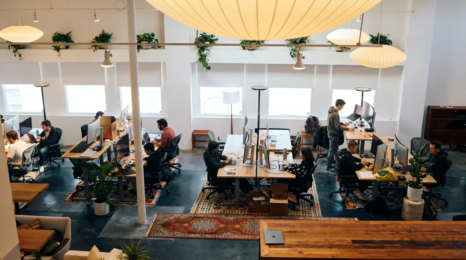 Wide elevated view of a warm studio office: glowing paper lanterns, a rail of trailing plants, oak desk pods and vintage rugs layered across the floor